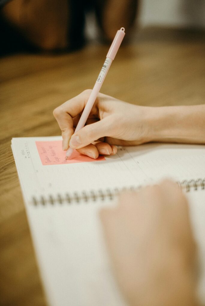 pexels-photo-3202029-3202029 Close-up of a person writing in a notebook with a pink pen on a wooden desk.